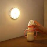 Motion Sensor LED night light wall-mounted round lamp glowing warm light in nursery beside baby bottle, battery-powered ABS